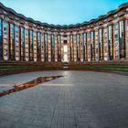 Ein Halbkreis aus hohen, vertikal strukturierten Gebäuden mit vielen Fenstern umgibt einen leeren, gepflasterten Platz.