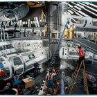 Ein Filmset einer Raumstation, mit technisch aussehenden Röhren, Leitern, Menschen in Arbeitskleidung und viel Ausrüstung.