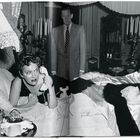 Eine Frau liegt in einem luxuriösen Raum auf einem Bett, hält einen Telefonhörer. Ein Mann steht im Hintergrund.