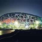 Eine beleuchtete Stadionstruktur mit verschlungenen, gitterartigen Streben, umgeben von Bäumen und Straßenlampen.