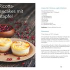 Mini-Ricotta-Cheesecakes mit Granatapfel. Zwei kleine Kuchen mit Mandeln. Granatapfelkerne in einer Schale im Hintergrund.