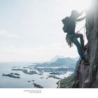 Der Text lautet: "Niels Pelzken in Lofoten, Norway, with Svolvær in the background". Ein Kletterer an einer steilen Wand mit weitem Blick aufs Meer.