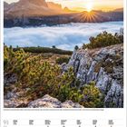 Monate und Tage in Deutsch, Sonnenaufgang über Berglandschaft, nebelige Täler, grünes Gebüsch und Felsen im Vordergrund.