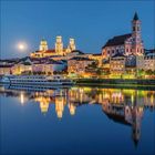 Kein relevanter Text. Abendliche Stadtansicht mit beleuchteten Gebäuden, Kirche, Fluss und Boot, alles spiegelnd im Wasser.