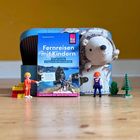 Der Text auf einem Buch lautet: "Fernreisen mit Kindern: Mit der Familie entspannt die Welt entdecken." Spielzeugfiguren und ein Plüschigel stehen daneben.