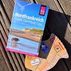 "Nordfrankreich: Picardie, Côte d'Opale, Calais." Reiseführer auf Holzhintergrund, daneben Deko in Postkartenform mit Aufschriften.