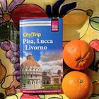 "CityTrip Pisa, Lucca, Livorno" Reiseführer auf einem bunten Tisch mit zwei Orangen daneben.