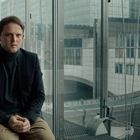 Ein Mann in dunkler Kleidung sitzt auf einem Stuhl in einem verglasten Büro, im Hintergrund moderne Gebäude.