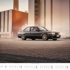 August. Wochentage in zwei Reihen; 1 beginnt am Sonntag. Schwarzer BMW E30 vor industrieller Kulisse bei Sonnenuntergang.
