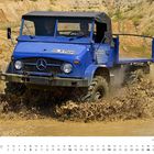 „DN N 950 H“ auf dem Nummernschild eines blauen Unimogs, der durch Matsch fährt, umgeben von sandigen Hügeln.