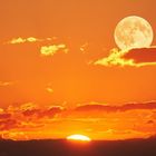 Sonnenuntergang: Der orange Himmel zeigt die Sonne, die hinter den Wolken versinkt, und einen Vollmond darüber.