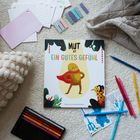 "Mut ist ein gutes Gefühl" auf Kinderbuch-Cover mit fröhlichem Charakter. Umgebung mit Decken und bunten Stiften.