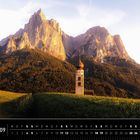 Kalender: September, Dolomiten. Ein Kirchturm auf grünem Feld vor imposanten, beleuchteten Bergen.