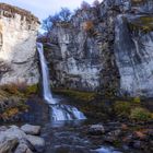 Ein hoher Wasserfall stürzt von einer Felswand in einen Fluss. Felsen und bunte Bäume umgeben die Szene.