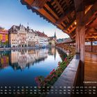 "Kapellbrücke in Luzern 9". Eine überdachte Holzbrücke mit Blumen führt über einen Fluss, Altstadt im Hintergrund.