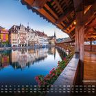 Kapellbrücke in Luzern 9. Ein hölzerner, überdachter Steg mit roten Blumen führt über einen Fluss; dahinter Häuserreihe.