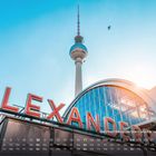 "Alexanderplatz" in großen roten Buchstaben. Fernsehturm vor blauem Himmel, Glasfassade erstrahlt im Sonnenlicht.