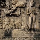 Steinrelief mit zwei Figuren, einer sitzenden und einer stehenden. Beide sind von Pflanzen und Ornamenten umgeben.