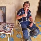 Ein Mann in Motorradkleidung sitzt lächelnd mit einem Glas Tee auf einem Stuhl. Neben ihm steht ein Tisch mit Teekanne.