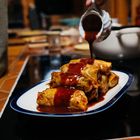 Eine Hand gießt rote Soße über gerollte Speisen auf einem weißen Teller in einer Küche mit warmen Holztönen.