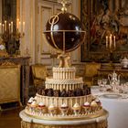 Kugel mit "N O S" Kompass, aufwendig dekoriert, steht auf einer mehrstufigen, mit Gebäck verzierten Torte in elegantem Raum.