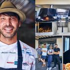 Ein Mann mit Hut lächelt, während auf seiner Jacke mehrere Firmenlogos zu sehen sind. Daneben zwei Holzöfen und eine kleine Gruppe von vier Männern.