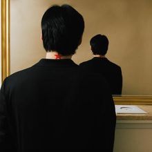 Kein Text im Bild. Eine Person steht vor einem Spiegel, vom Rücken aus gesehen, mit einem roten Fleck am Nacken.