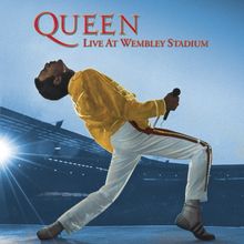 "QUEEN LIVE AT WEMBLEY STADIUM" in roter Schrift oben. Ein Performer in gelber Jacke und weißer Hose, im Stadion.