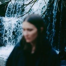 Kein sichtbarer Text. Eine unscharfe Person steht vor einem Wasserfall, umgeben von Bäumen und Wasserströmen.