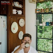 Text: "Niall Horan Dinner Party". Eine Person trinkt aus einer Tasse in einer rustikalen Küche mit Geschirr an den Wänden und einem Fenster.