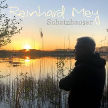 Text: "Reinhard Mey Schatzhauser". Eine Silhouette steht vor einem See bei Sonnenuntergang, umgeben von Schilf.