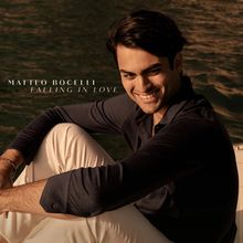 MATTEO BOCELLI FALLING IN LOVE. Ein lächelnder Mann sitzt entspannt an einem sonnigen Ort mit Wasser im Hintergrund.