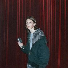 Eine Person mit langen Haaren hält ein Mikrofon und trägt kariertes Hemd und Jacke. Hintergrund: rote Vorhänge.