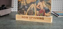 "NOW SPINNING" steht auf einem hölzernen Ständer von Crosley. Dahinter ist ein Gemälde von mehreren Personen zu sehen.