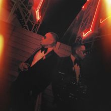 Zwei Männer in eleganten Anzügen und Sonnenbrillen stehen im Dunkeln vor neonrotem Licht, das ein stilvolles Ambiente schafft.