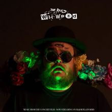 "The Real Will Wood" steht oben. Ein Mann mit Bart, Blumenkranz und großer Brille, grün beleuchtet, schaut ernst.