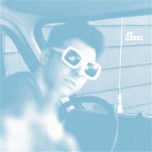 "N° 235" ist auf einem blauen Hintergrund zu sehen. Eine unscharfe Figur mit großer Sonnenbrille sitzt im Auto.