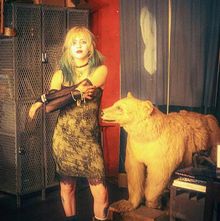 Keine erkennbaren Texte. Eine Person mit Tattoos steht neben einem ausgestopften Bären in einem rustikalen Raum.