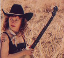 Frau mit Hut und Banjo in trockener Landschaft, Tätowierung mit Cowboyhut und Noten auf dem Arm.