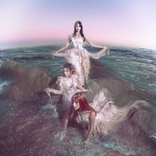 Drei Frauen in fließenden Kleidern stehen und knien auf Felsen im Meer unter einem pastellfarbenen Himmel.