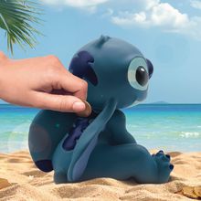 Eine Hand steckt eine Münze in ein blaues Wesen in Tierform, das am Sandstrand sitzt, mit Meer und Palmen im Hintergrund.