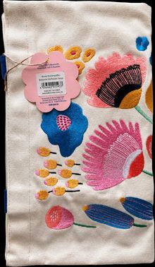 "Bunte Blumengrüße", bestickter Stoffbeutel in Beige mit farbenfrohen Blumenmustern und einem rosa Etikett in Blütenform.