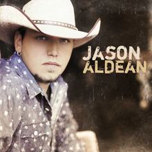 „JASON ALDEAN“ in groben Buchstaben. Person mit Cowboyhut, trägt ein gemustertes Hemd, schaut direkt nach vorn.