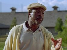 Ein Mann mit Mütze und hellem Hemd steht draußen, zeigt mit der Hand. Im Hintergrund ist eine Mauer mit Türmen.