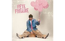Text: "FÊTE FORAINE". Ein Mann in bunter Kleidung sitzt lächelnd mit rosa Luftballons.