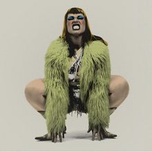 Keine sichtbaren Texte. Eine Person in grüner Felljacke, blauem Lidschatten, aggressiver Pose.