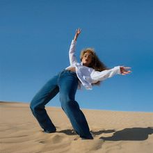 Eine Person in lockerer Pose auf einer Sanddüne, trägt weites weißes Hemd und Jeans, strahlend blauer Himmel im Hintergrund.
