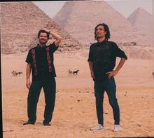 Zwei Männer stehen vor den Pyramiden von Gizeh, umgeben von Sand. Im Hintergrund sind Kamele und Gespanne zu sehen.