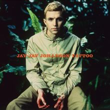 „JAY-JAY JOHANSON TATTOO“ in orange. Ein Mann in hellem Oberteil sitzt in üppiger Vegetation.