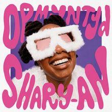 „OPMNWTD Shary-An“. Eine fröhliche Person mit großen, flauschigen Sonnenbrillen und Lächeln vor einem lila Hintergrund.